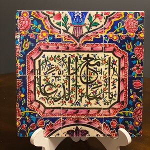May include: A ceramic tile with a colorful floral design and Arabic calligraphy in a white frame. The tile is on a white stand.