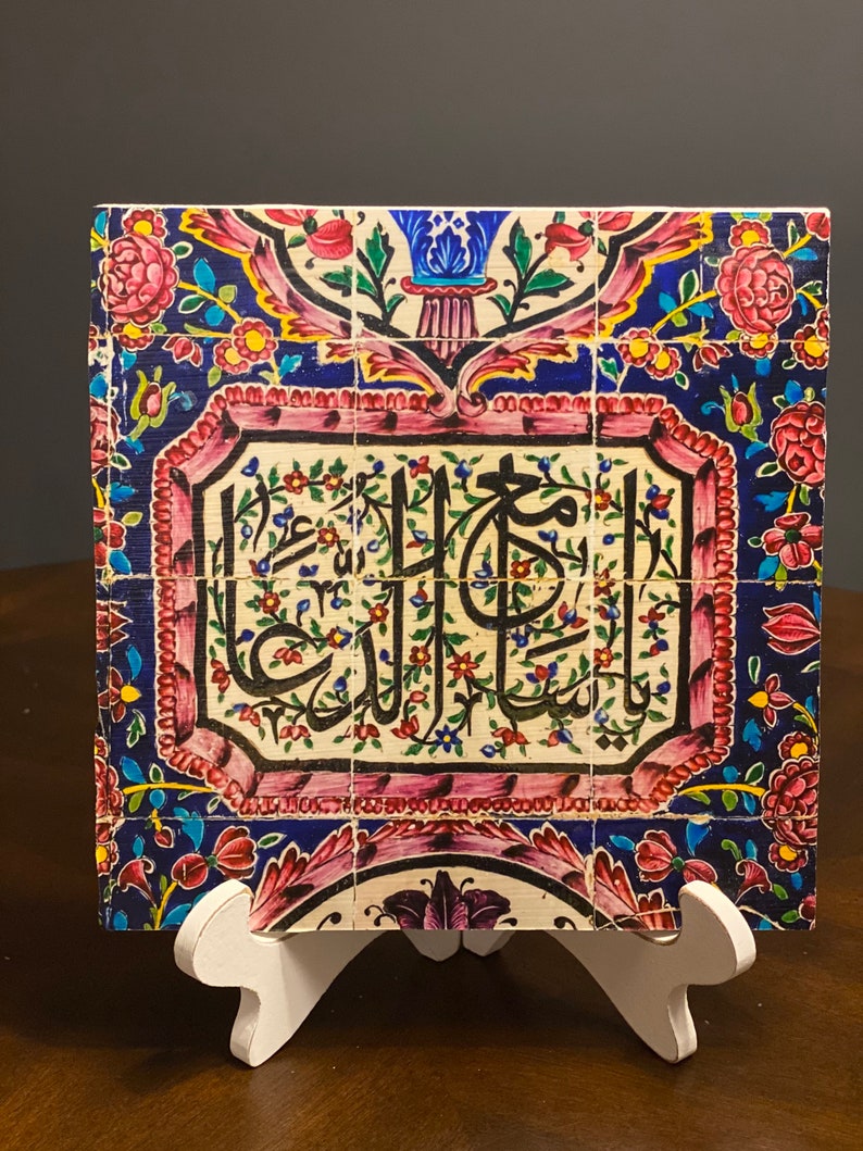 May include: A ceramic tile with a colorful floral design and Arabic calligraphy in a white frame. The tile is on a white stand.