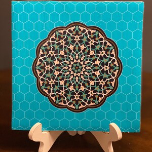 May include: A turquoise blue square tile with a hexagonal pattern. The tile features a large, intricate mandala design in shades of black, white, green, brown, and gold. The mandala is surrounded by a white border.