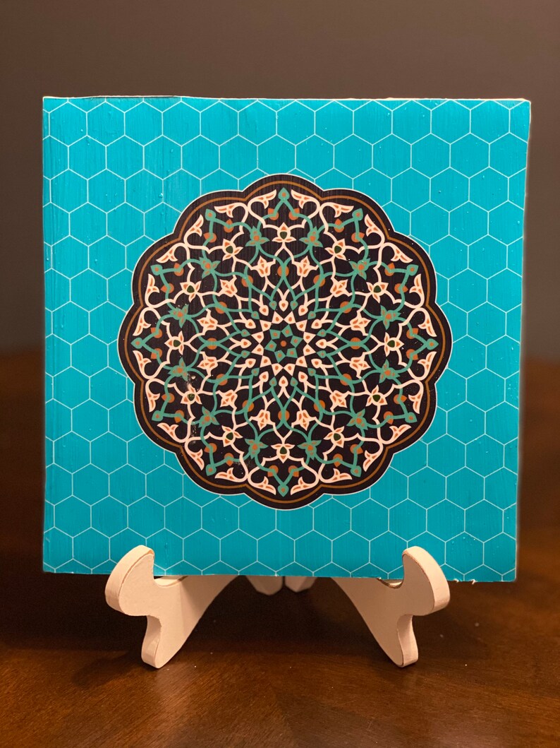 May include: A turquoise blue square tile with a hexagonal pattern. The tile features a large, intricate mandala design in shades of black, white, green, brown, and gold. The mandala is surrounded by a white border.