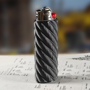 May include: A black and silver lighter with a textured, rope-like design. The lighter has a silver metal top with a red ignition button. The lighter is resting on a sheet of paper with printed text.