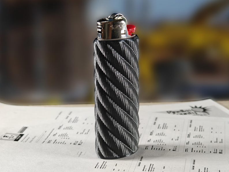 May include: A black and silver lighter with a textured, rope-like design. The lighter has a silver metal top with a red ignition button. The lighter is resting on a sheet of paper with printed text.