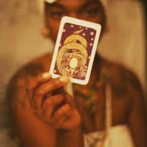 Puede incluir: Una carta de tarot con una ilustración detallada de una serpiente dorada y un óvalo blanco central, sostenida frente a una persona. La carta tiene un borde blanco y un fondo morado con pequeños puntos blancos.