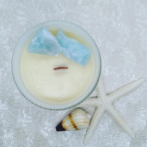 Puede incluir: Una vela blanca en un recipiente de vidrio con dos cristales azul claro en la parte superior. Una concha y una estrella de mar blanca están al lado de la vela sobre una superficie blanca estampada. La vela tiene una mecha de madera.