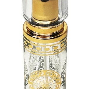 May include: A clear glass perfume bottle with gold accents. The bottle has a clear, rectangular cap and a gold-colored top. The body of the bottle is decorated with gold filigree designs. The bottle is designed to hold perfume or essential oils.