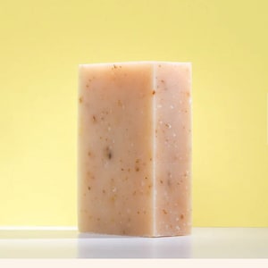 Puede incluir: Una barra de jabón rectangular de color rosa claro con pequeñas motas marrones en todo. El jabón está sobre una superficie amarilla.