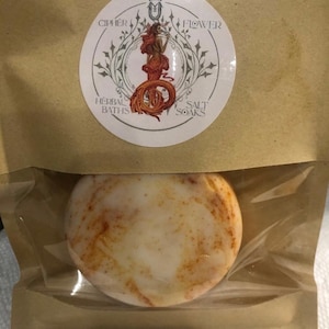 Puede incluir: Un baño de burbujas redondo y blanco con motas marrones, empaquetado en una bolsa de papel marrón con una etiqueta que dice "Cipher Flower Herbal Baths Salt Soaks".