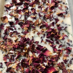 以下が含まれることがあります： 白い石鹸のベースに、乾燥したバラの花びらと薄い茶色の粉末がまぶされています。石鹸のベースは透明な長方形の容器に入っています。