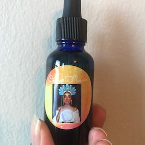 Puede incluir: Una pequeña botella de vidrio oscuro con un gotero azul. La botella tiene una etiqueta redonda con una ilustración colorida de una mujer con una corona y sosteniendo una bola de cristal. La etiqueta dice "Chaser Flower".