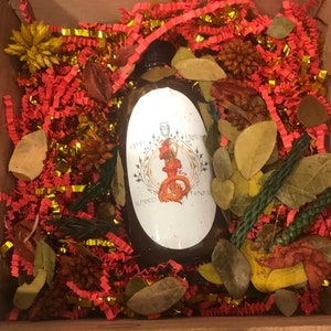 Puede incluir: Una botella de vidrio con un líquido marrón en su interior. La botella tiene una etiqueta blanca con un diseño de sirena y el texto "Blessed" y "Divine". La botella está rodeada de papel arrugado rojo y dorado.