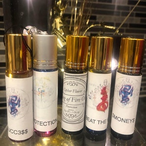 Puede incluir: Cinco pequeños frascos de vidrio con tapas doradas y etiquetas. Las etiquetas tienen diferentes diseños y textos, que incluyen "Cipher Flower", "King of Fire Oil", "Protection", "Beat the" y "Moneys".