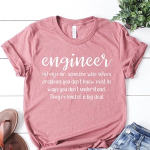 May include: A pink t-shirt with the word "engineer" printed on the front. The definition of the word is printed below: "(n) enja nir: someone who solves problems you don't know exist in ways you don't understand. They're kind of a big deal."
