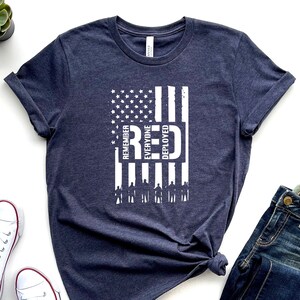 Könnte beinhalten: Ein marineblaues T-Shirt mit einem weißen, abgenutzten Design einer amerikanischen Flagge. Die Flagge trägt die vertikal geschriebenen Worte "Remember Everyone Deployed" und "RED".