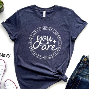 Könnte beinhalten: Heather-Marineblaues T-Shirt mit einem weißen Kreis-Motiv, auf dem "You are" steht, umgeben von den Wörtern "capable", "enough", "worthy", "loved", "kind" und "strong".