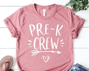 May include: A pink t-shirt with the text "PRE-K CREW" in white with an arrow and heart design. The shirt is laying on a white wooden surface with a pair of brown sandals, blue jeans, and a potted plant.