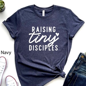 May include: Heather navy blue t-shirt with white text that reads "Raising tiny disciples" with a small heart symbol.