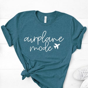 May include: Teal t-shirt with the words "airplane mode" in white script and a small airplane graphic. The shirt is knotted at the bottom and is paired with white sneakers. Small potted succulents are placed around the shirt on a white wooden surface.