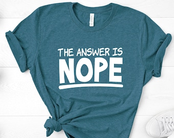 May include: A teal blue t-shirt with the text "THE ANSWER IS NOPE" in white lettering. The shirt is tied in a knot and laid on a white wooden surface.