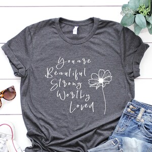 Könnte beinhalten: Ein dunkelgraues T-Shirt mit dem Text "You are Beautiful Strong Worthy Loved" und einer einfachen Blumengrafik.