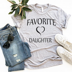 Puede incluir: Una camiseta gris con el texto "FAVORITE" y un s&iacute;mbolo de coraz&oacute;n sobre el texto "DAUGHTER". La camiseta est&aacute; colocada sobre una superficie blanca con un par de pantalones vaqueros, gafas de sol y zapatos blancos.
