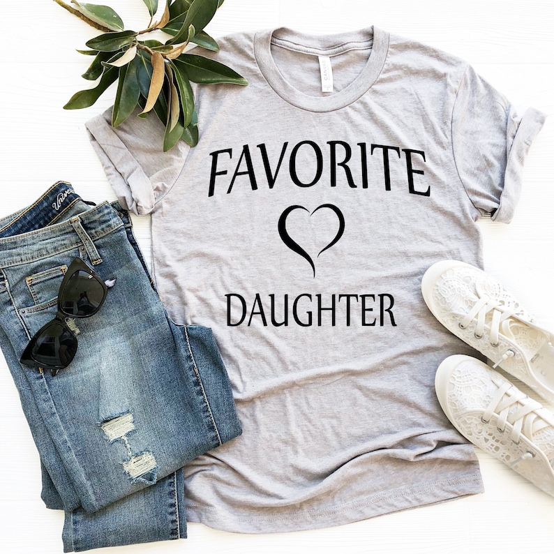 Puede incluir: Una camiseta gris con el texto "FAVORITE" y un s&iacute;mbolo de coraz&oacute;n sobre el texto "DAUGHTER". La camiseta est&aacute; colocada sobre una superficie blanca con un par de pantalones vaqueros, gafas de sol y zapatos blancos.