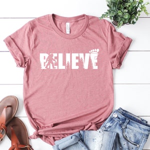 May include: A pink t-shirt with the words "BELIEVE" in white letters. The letters are made up of a silhouette of a Bigfoot, trees, and a footprint.