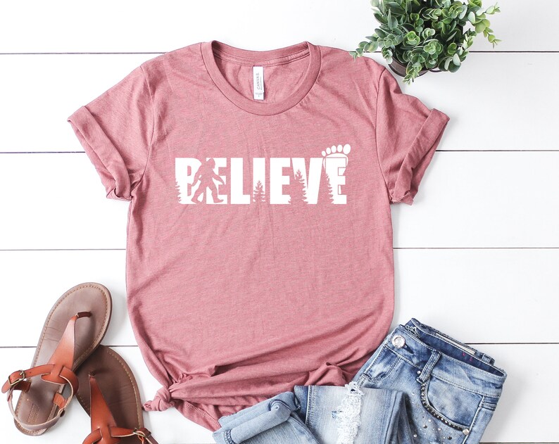 May include: A pink t-shirt with the words "BELIEVE" in white letters. The letters are made up of a silhouette of a Bigfoot, trees, and a footprint.