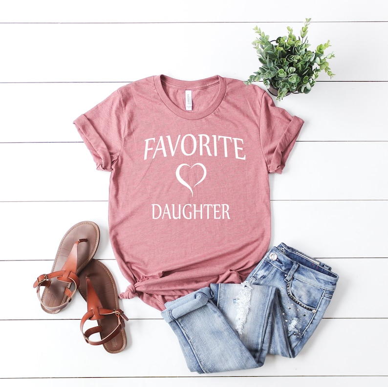 Puede incluir: Una camiseta color malva con el texto "FAVORITE" y un s&iacute;mbolo de coraz&oacute;n sobre la palabra "DAUGHTER" en blanco.