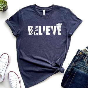 May include: A dark blue t-shirt with a white graphic that says "BELIEVE" with a silhouette of Bigfoot and a footprint.