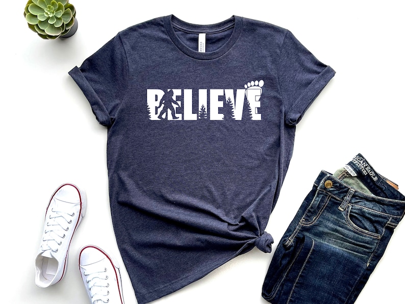 May include: A dark blue t-shirt with a white graphic that says "BELIEVE" with a silhouette of Bigfoot and a footprint.