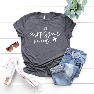 May include: Dark gray t-shirt with the words "airplane mode" in white script and a small airplane graphic. The shirt is paired with blue jeans, white sneakers, and sunglasses, suggesting a travel theme.