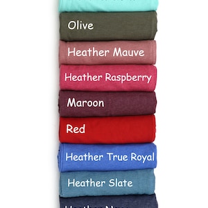 May include: A stack of folded t-shirts in various colors, including white, pink, olive, maroon, red, blue, and gray. The t-shirts are labeled with their color names.