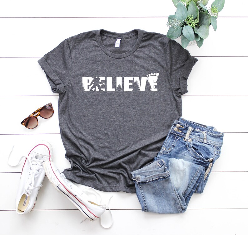 May include: A dark gray t-shirt with a white graphic of a Bigfoot and a footprint, with the word "BELIEVE" spelled out in the graphic. The shirt is laid out on a white wooden surface with a pair of white sneakers, a pair of sunglasses, and a pair of blue jeans.