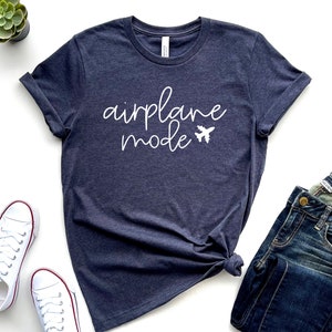 May include: Navy blue t-shirt with the words "airplane mode" in white script and a small airplane graphic. The shirt is paired with blue jeans and white sneakers. A small green plant sits in a black pot.