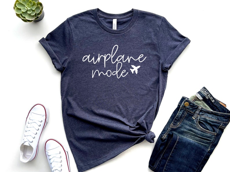 May include: Navy blue t-shirt with the words "airplane mode" in white script and a small airplane graphic. The shirt is paired with blue jeans and white sneakers. A small green plant sits in a black pot.