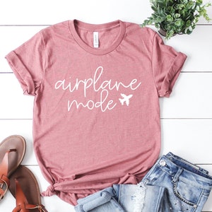May include: A dusty rose t-shirt with the words "airplane mode" in white script and a small airplane graphic. The shirt is paired with blue jeans and brown sandals, suggesting a travel theme.