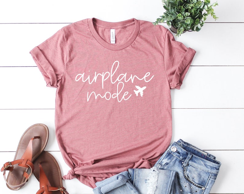 May include: A dusty rose t-shirt with the words "airplane mode" in white script and a small airplane graphic. The shirt is paired with blue jeans and brown sandals, suggesting a travel theme.