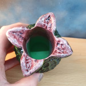 Może przedstawiać: Unikalny, ręcznie robiony ceramiczny wazon z motywem kwiatu Demogorgona. Wazon ma zieloną podstawę i teksturowaną, ciemnozieloną powierzchnię zewnętrzną. Płatki kwiat&oacute;w są głęboko czerwone z białymi akcentami, a wnętrze wypełnione jest zielonym płynem.