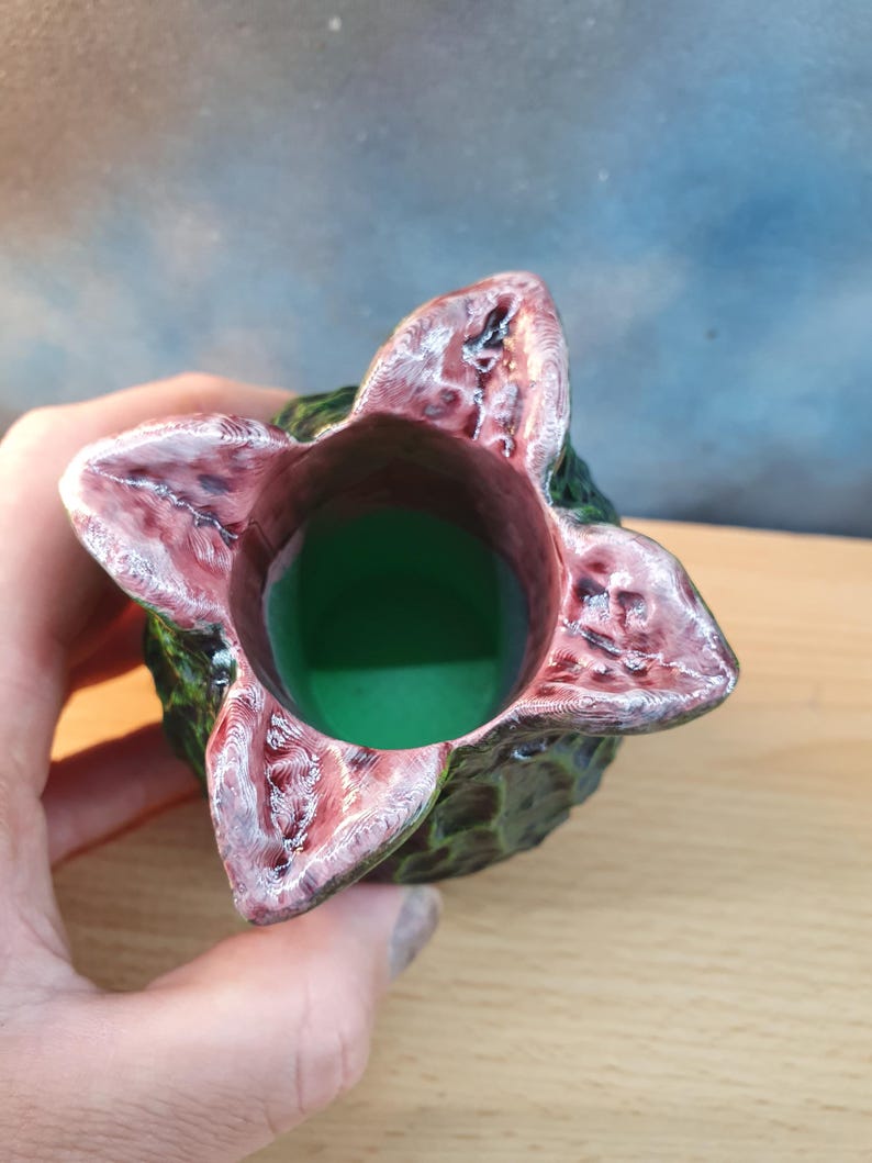Może przedstawiać: Unikalny, ręcznie robiony ceramiczny wazon z motywem kwiatu Demogorgona. Wazon ma zieloną podstawę i teksturowaną, ciemnozieloną powierzchnię zewnętrzną. Płatki kwiat&oacute;w są głęboko czerwone z białymi akcentami, a wnętrze wypełnione jest zielonym płynem.