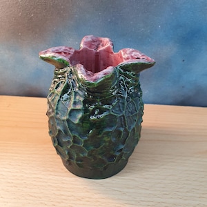 May include: A green and black textured vase with a red interior. The vase has a unique, organic shape with a flared top. The exterior has a rough, bumpy texture, and the interior is smooth. The vase is sitting on a wooden surface.