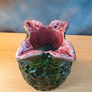 Może przedstawiać: Ręcznie wykonany ceramiczny pojemnik o unikalnym, organicznym kształcie. Zewnętrzna strona jest teksturowana, ciemnozielona, ​​a wnętrze i obręcz są bogate w czerwono-fioletowy. Otwarty wierzch odsłania ciemnozielone wnętrze.