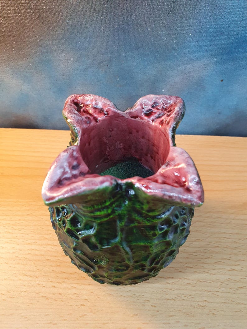 Może przedstawiać: Ręcznie wykonany ceramiczny pojemnik o unikalnym, organicznym kształcie. Zewnętrzna strona jest teksturowana, ciemnozielona, ​​a wnętrze i obręcz są bogate w czerwono-fioletowy. Otwarty wierzch odsłania ciemnozielone wnętrze.