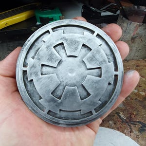 Op de afbeelding: Een rond, zilverkleurig metalen embleem met een gestileerd sterrendesign. Het embleem heeft een verweerde, verweerde uitstraling met een grijze patina.