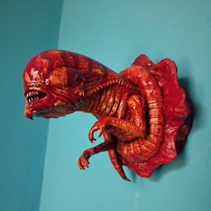 May include: A detailed, red-orange sculpture of an alien face and chest, mounted on a wall. The creature has a large head, sharp teeth, and detailed musculature. The sculpture is affixed to a red, textured base, with a blue wall in the background.