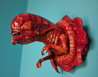 May include: A detailed, red-orange sculpture of an alien face and chest, mounted on a wall. The creature has a large head, sharp teeth, and detailed musculature. The sculpture is affixed to a red, textured base, with a blue wall in the background.