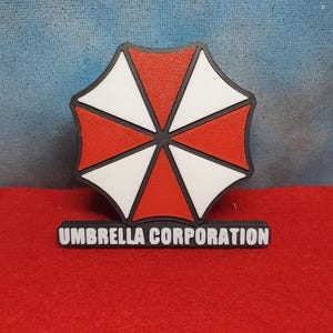 Könnte beinhalten: Ein 3D-gedrucktes Schirmlogo in Schwarz, Weiß und Rot mit dem Text "Umbrella Corporation" darunter. Das Logo ist ein roter und weißer Schirm mit schwarzem Umriss.