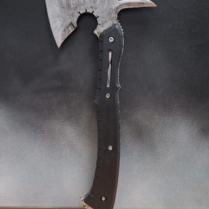 Pode incluir: Um machado de metal desgastado com um cabo preto. A cabeça do machado tem uma lâmina curva, uma extremidade pontiaguda e um recorte circular. O cabo é texturizado e fixado com rebites. O machado é projetado para utilidade.