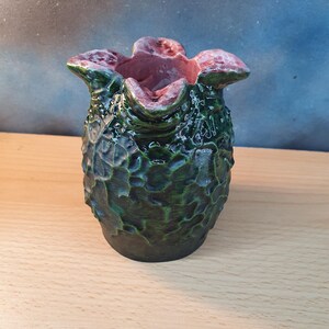 Może przedstawiać: Ręcznie wykonany ceramiczny wazon o unikalnej, teksturowanej powierzchni. Wazon ma ciemnozielony zewnętrzny wygląd z jaśniejszymi zielonymi akcentami i czerwonor&oacute;żowym wnętrzem. Projekt jest organiczny i rzeźbiarski, przypominający stylizowany kształt kwiatu lub rośliny.