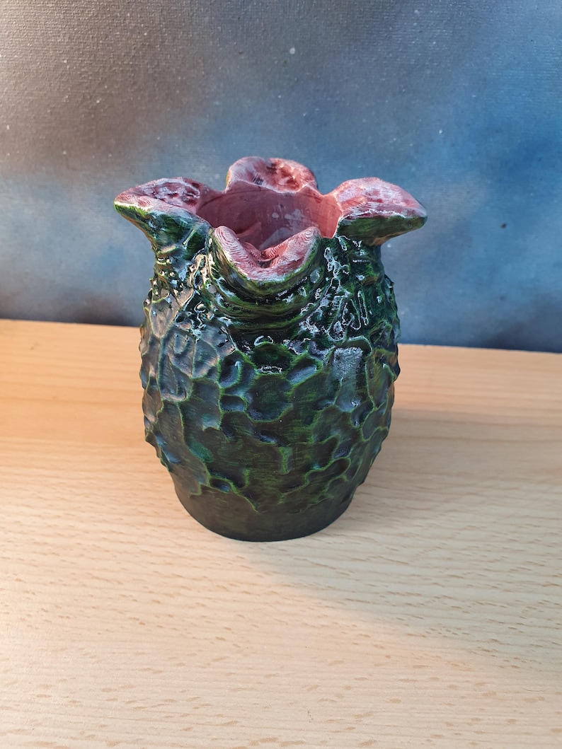 Może przedstawiać: Ręcznie wykonany ceramiczny wazon o unikalnej, teksturowanej powierzchni. Wazon ma ciemnozielony zewnętrzny wygląd z jaśniejszymi zielonymi akcentami i czerwonor&oacute;żowym wnętrzem. Projekt jest organiczny i rzeźbiarski, przypominający stylizowany kształt kwiatu lub rośliny.