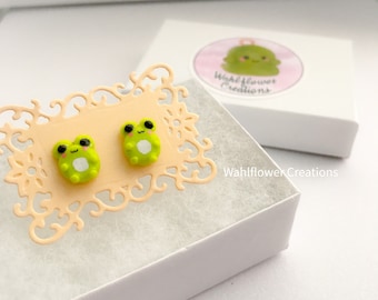 Può includere: Un paio di orecchini a bottone a forma di rana verde in una scatola bianca con un logo rosa e verde che dice "Wahlflower Creations".